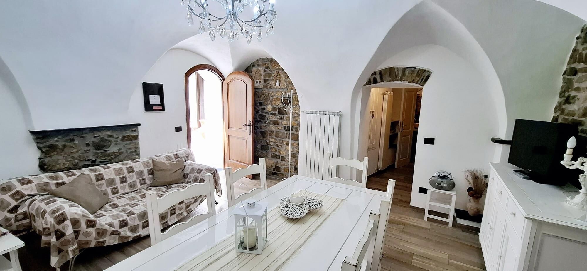 Un salon chaleureux avec un canapé, une table à manger et un lustre. Les murs sont en pierre et les portes en bois.