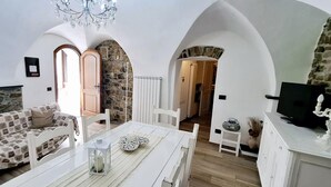 A cozy living room with a sofa, a dining table, and a chandelier. There are stone walls and wooden doors. - "A TASTE OF PROVENCE" Among Olive Trees and the Sea (Terzorio)