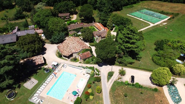 Une vue aérienne d’une propriété dotée d’une piscine, de courts de tennis et de plusieurs maisons entourées de verdure.