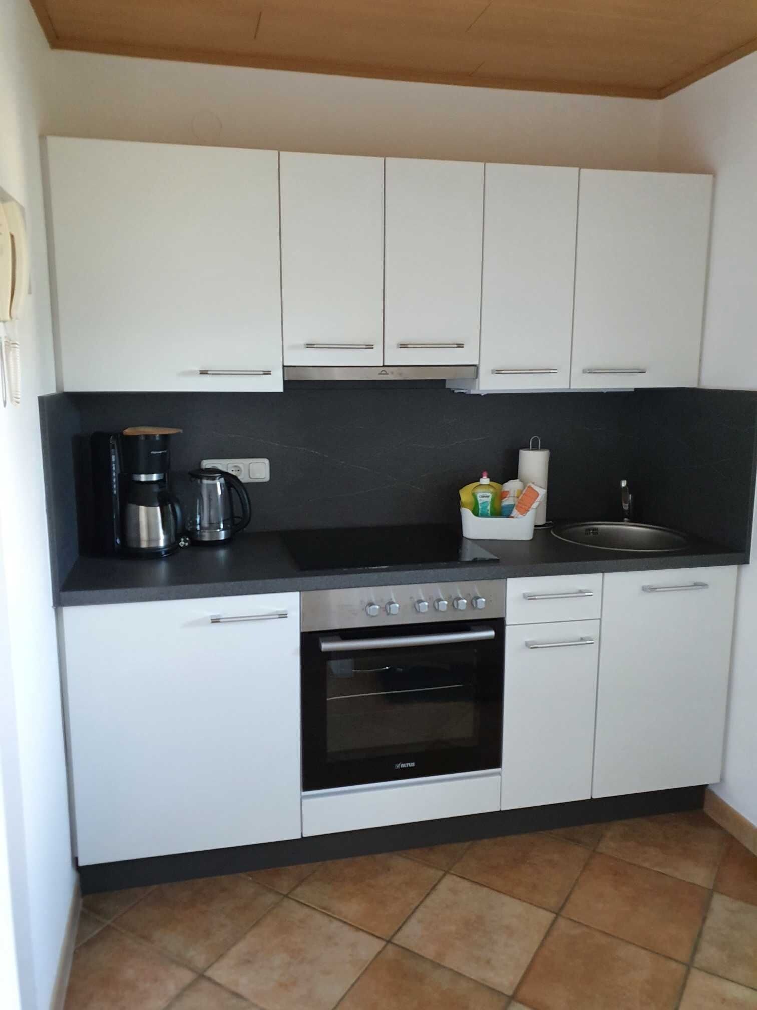 A compact kitchen with white cabinets, a black countertop, a coffee maker, a kettle, and a sink.