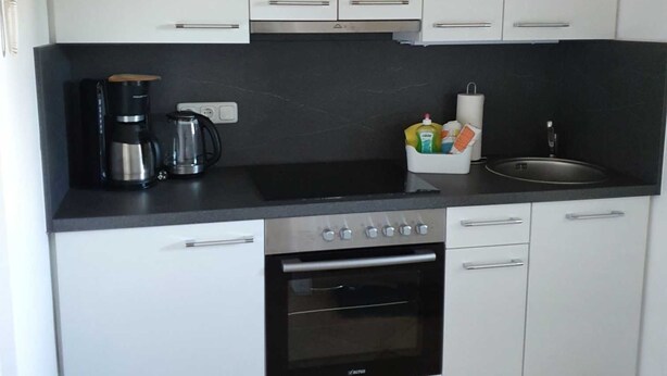 A compact kitchen with white cabinets, a black countertop, a coffee maker, a kettle, and a sink.
