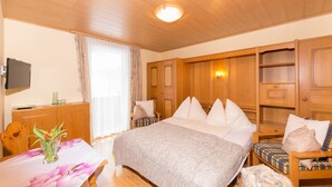 A bedroom with a bed, a wooden dresser, a chair, a table with a vase, and a television on the wall. - Apartment garden view - Apartment house Roland (Strobl)