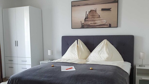 A bedroom with a bed, a nightstand, a white wardrobe, and a painting of a pier. - Apartment garden view - Apartment house Roland (Strobl)