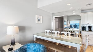 Ein modernes Zimmer mit einem Glastisch, weißen Stühlen, einem blauen Ottomanen mit Muster, einer Lampe und einem durchgehenden Durchgang zur Küche.