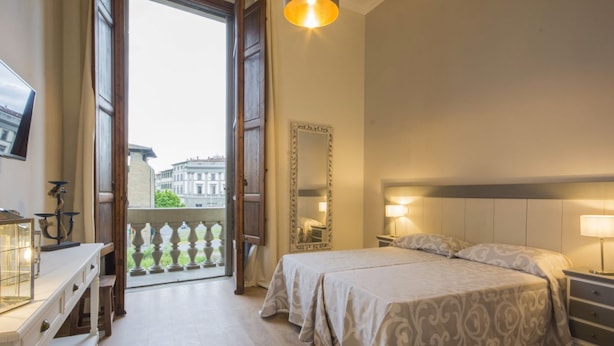 A bedroom with a bed, bedside tables, a mirror, a desk, and a window with a view of buildings.