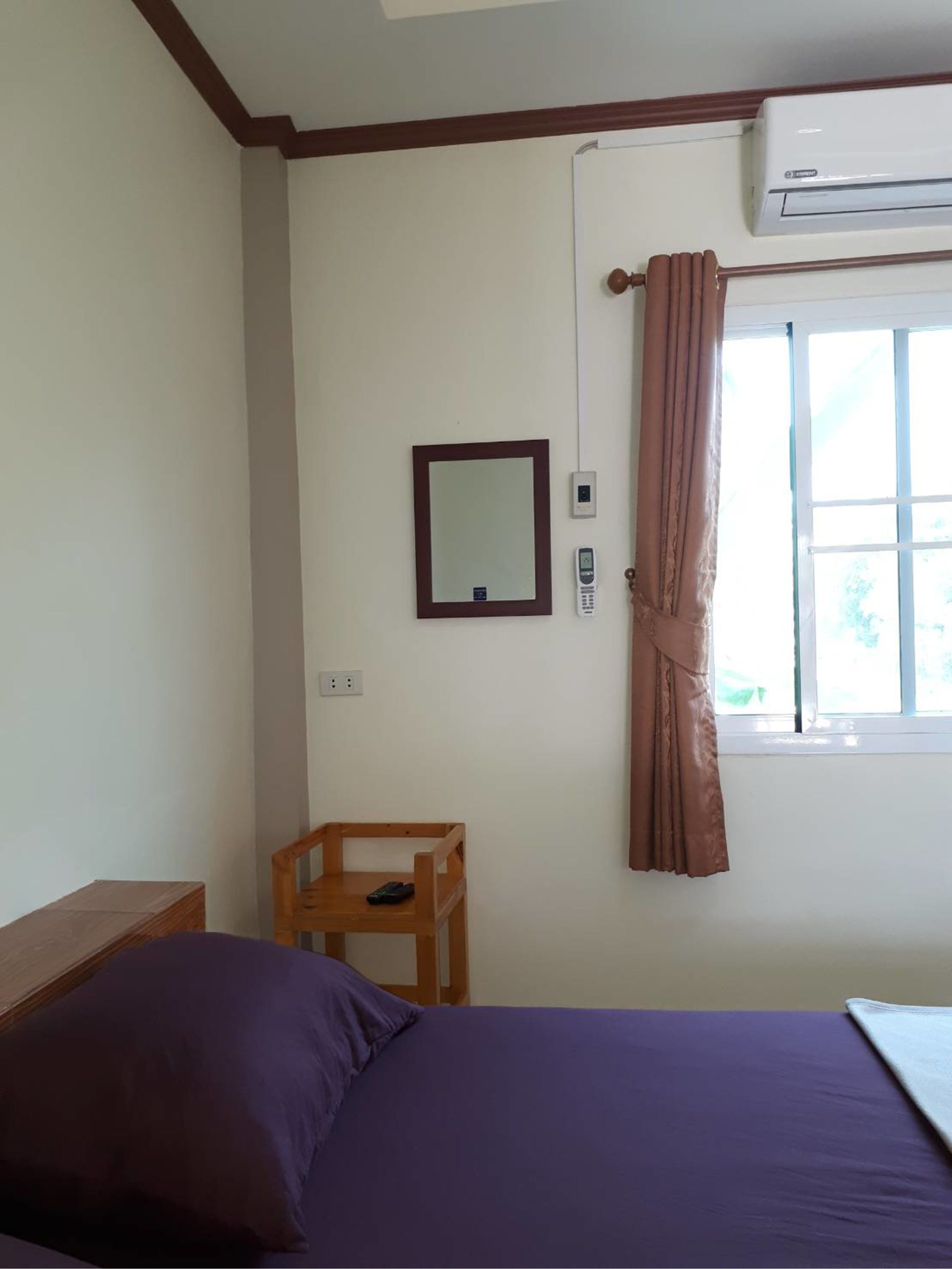 A bedroom with a bed, a wooden chair, a bedside table, a mirror, an air conditioning unit, and a window with curtains.