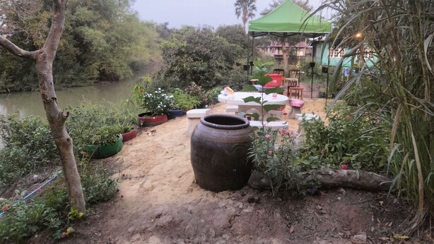 A riverside outdoor seating area with tables and chairs, surrounded by greenery and a large pot. - Dome Resort (Sena)