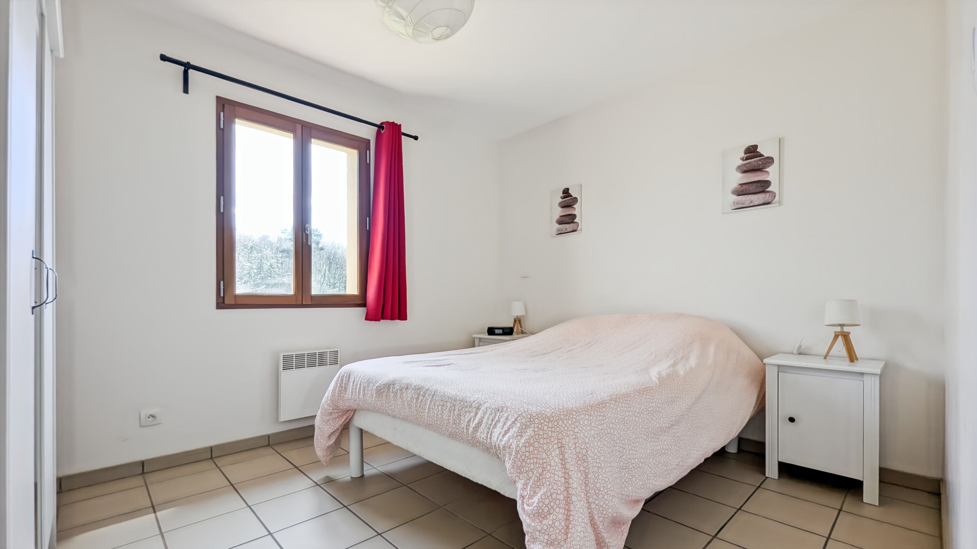 A bedroom with a bed, a window with curtains, a bedside table with a lamp, and a wall-mounted heater.