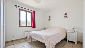 A bedroom with a bed, a window with curtains, a bedside table with a lamp, and a wall-mounted heater. - Little House with beautiful view in unique Holiday Resort **** in Dordogne-Lot (Gavaudun)