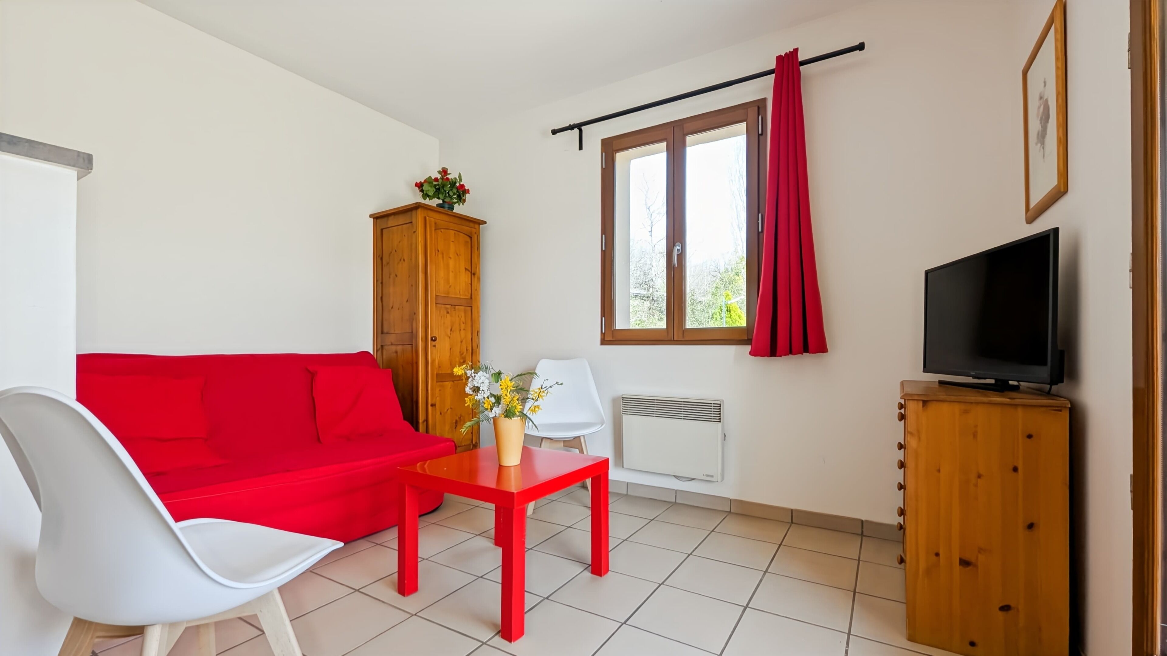 Une pièce avec un canapé rouge, une chaise blanche, une petite table ornée de fleurs, un téléviseur posé sur un meuble en bois et une fenêtre avec des rideaux.