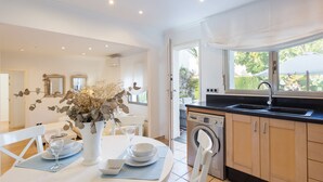 A modern kitchen with a built-in washing machine, a dining area with a white table set for two, and a view of the outdoors through a large window. - BOUGAINVILLEA HOUSE. COSTA VALENCIANA, COSTA BLANCA Pool, Garden, Beach, Villages. (Daimús)