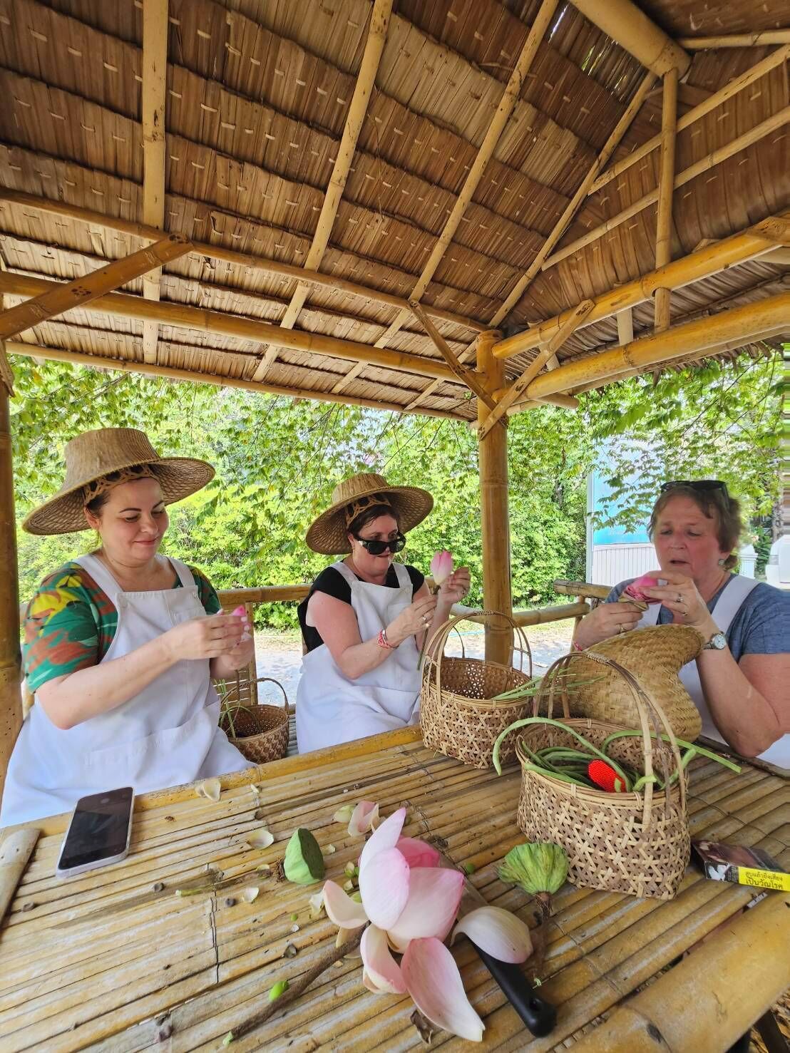 Three people working at a bamboo table with woven baskets, surrounded by various plants and tools.