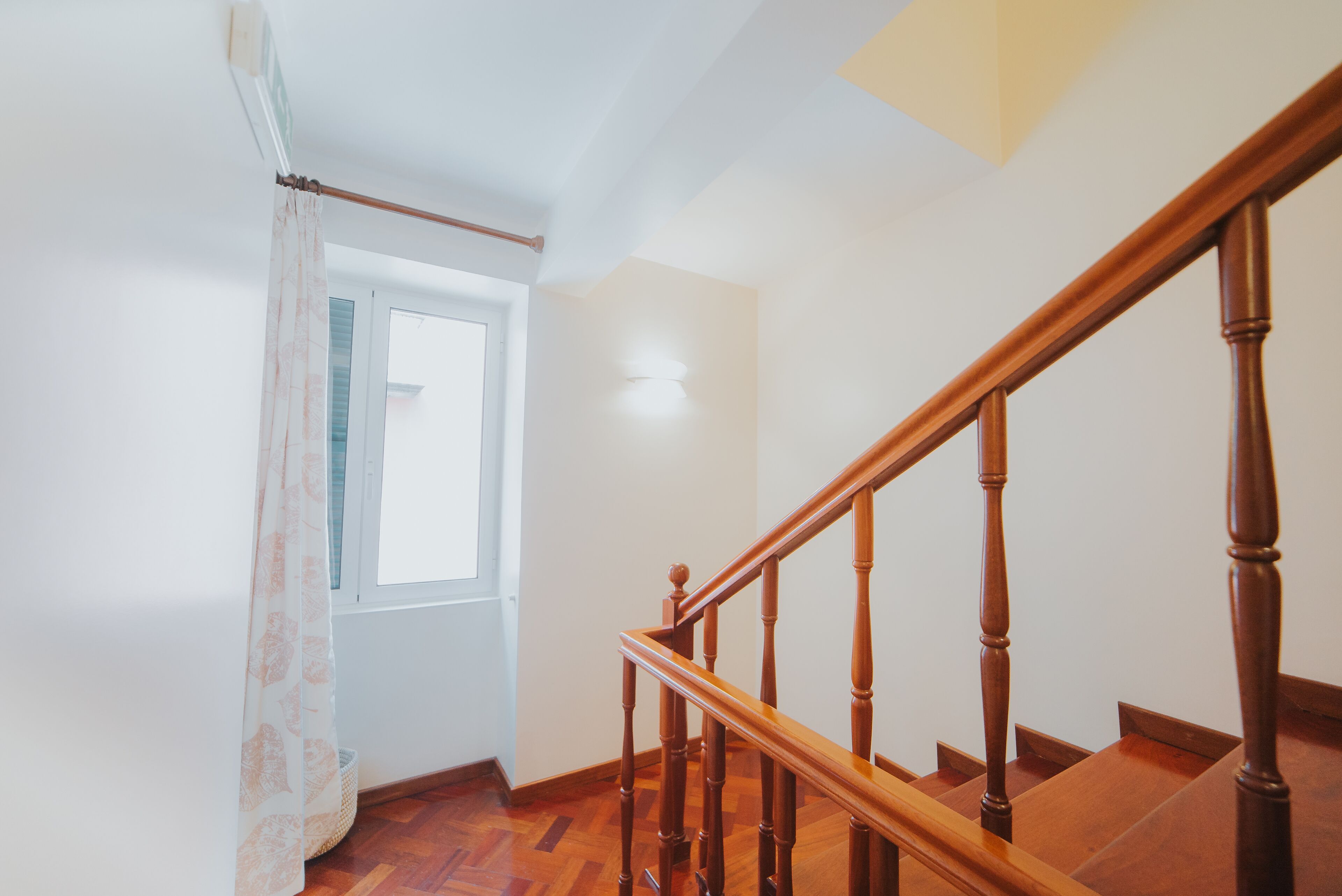 Eine Holztreppe mit Handlauf, ein Fenster mit Vorhang und eine Wandleuchte.
