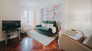 Ein Wohnzimmer mit einem grünen Sofa, einem weißen Sessel, einem Fernseher auf einem Schrank und floralem Wandpapier.