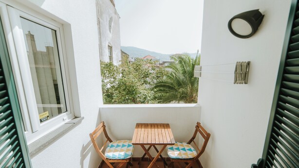 Ein kleiner Balkon mit Holztisch und zwei Stühlen, ein Fenster und Blick auf Grünflächen und Gebäude.