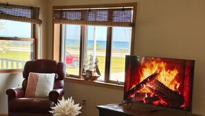 Ein gemütliches Wohnzimmer mit einem Ledersessel, einem Couchtisch mit einem Buch und einer Vase, einem Fernseher, der einen Kamin zeigt, und großen Fenstern mit Blick ins Freie.