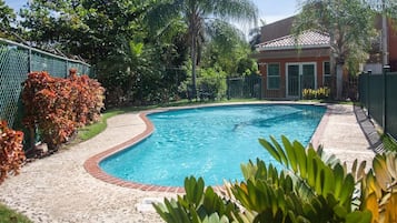 Una piscina con borde de ladrillo, rodeada por una valla, diversas plantas y árboles.