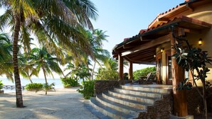 Een strandhuis met een stenen trap naar een houten veranda, palmbomen en uitzicht op zee.