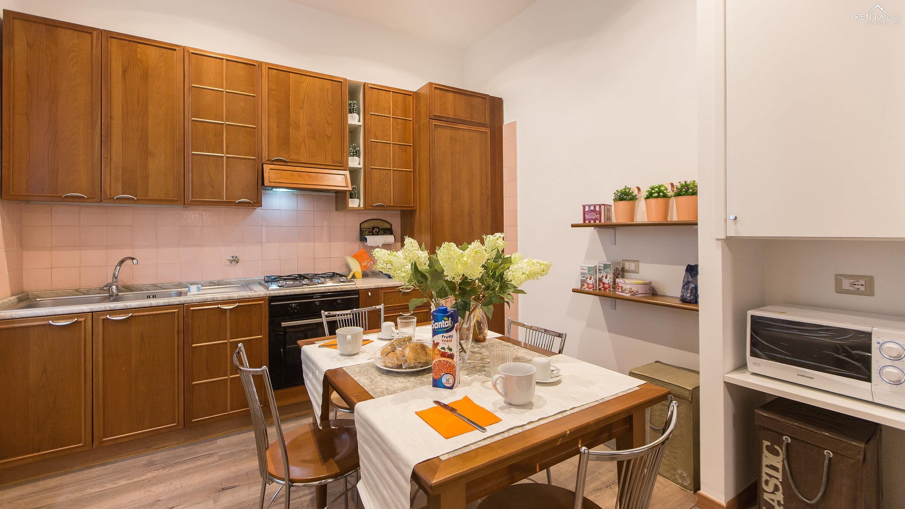 Une cuisine compacte avec des meubles en bois, une cuisinière à gaz et un four encastré. On y trouve une table à manger avec des chaises et un vase rempli de fleurs.