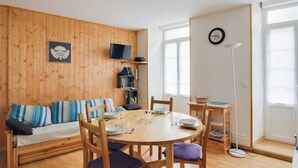 A cozy living room with a wooden floor, a sofa with striped cushions, a round dining table with chairs, a television mounted on the wall, and a large window with sheer curtains. - Central Apartment with 1 Bedroom, Ski Locker, Near Lift (Cauterets)
