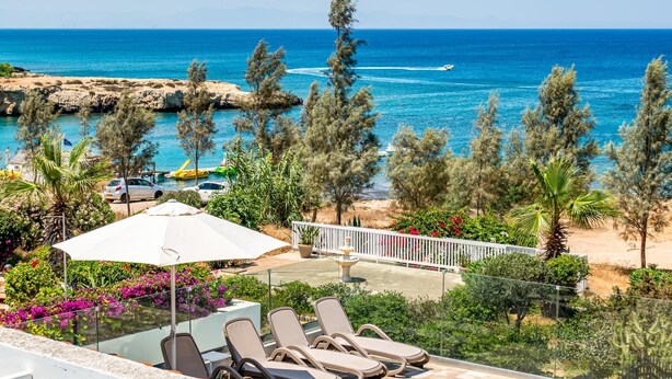 A balcony with lounge chairs and an umbrella overlooking a beach with clear blue water and rocky coastline. - Villa Aqua Marina by Ezoria Villas (Protaras)