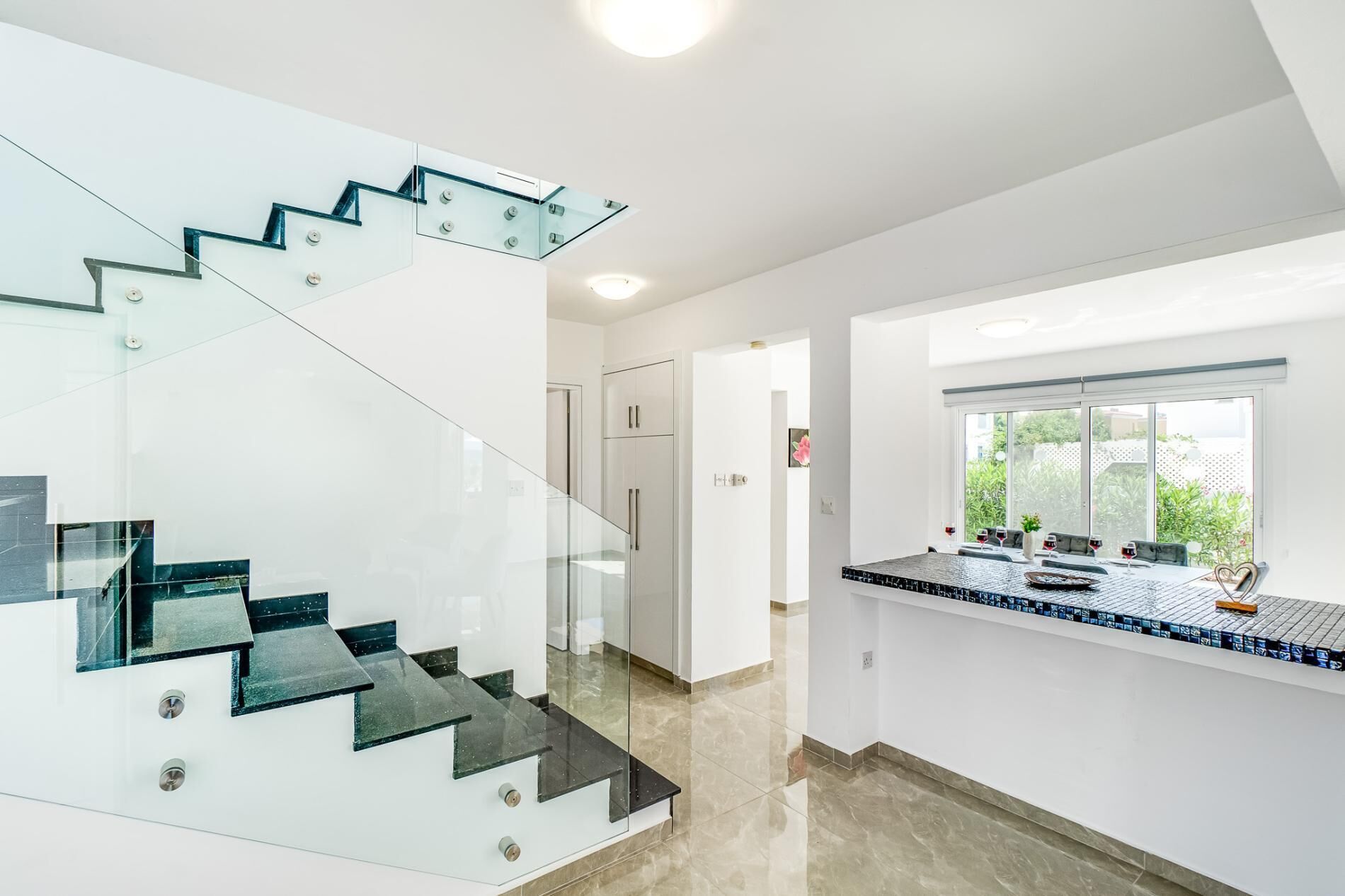 A modern interior with a glass staircase, a kitchen area with a tiled backsplash, and a view through a window.