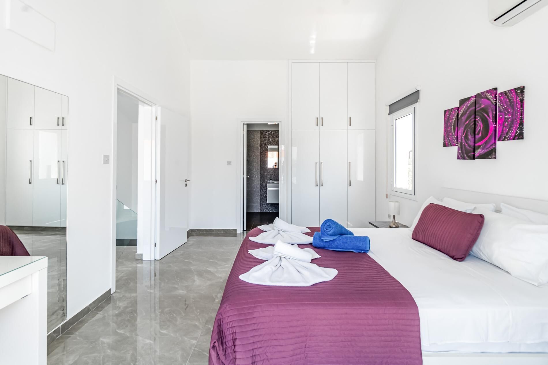 Une chambre d’hôtel moderne avec un grand lit, une literie blanche et violette, une salle de bain dotée d’une douche en verre et un couloir menant à une autre chambre.