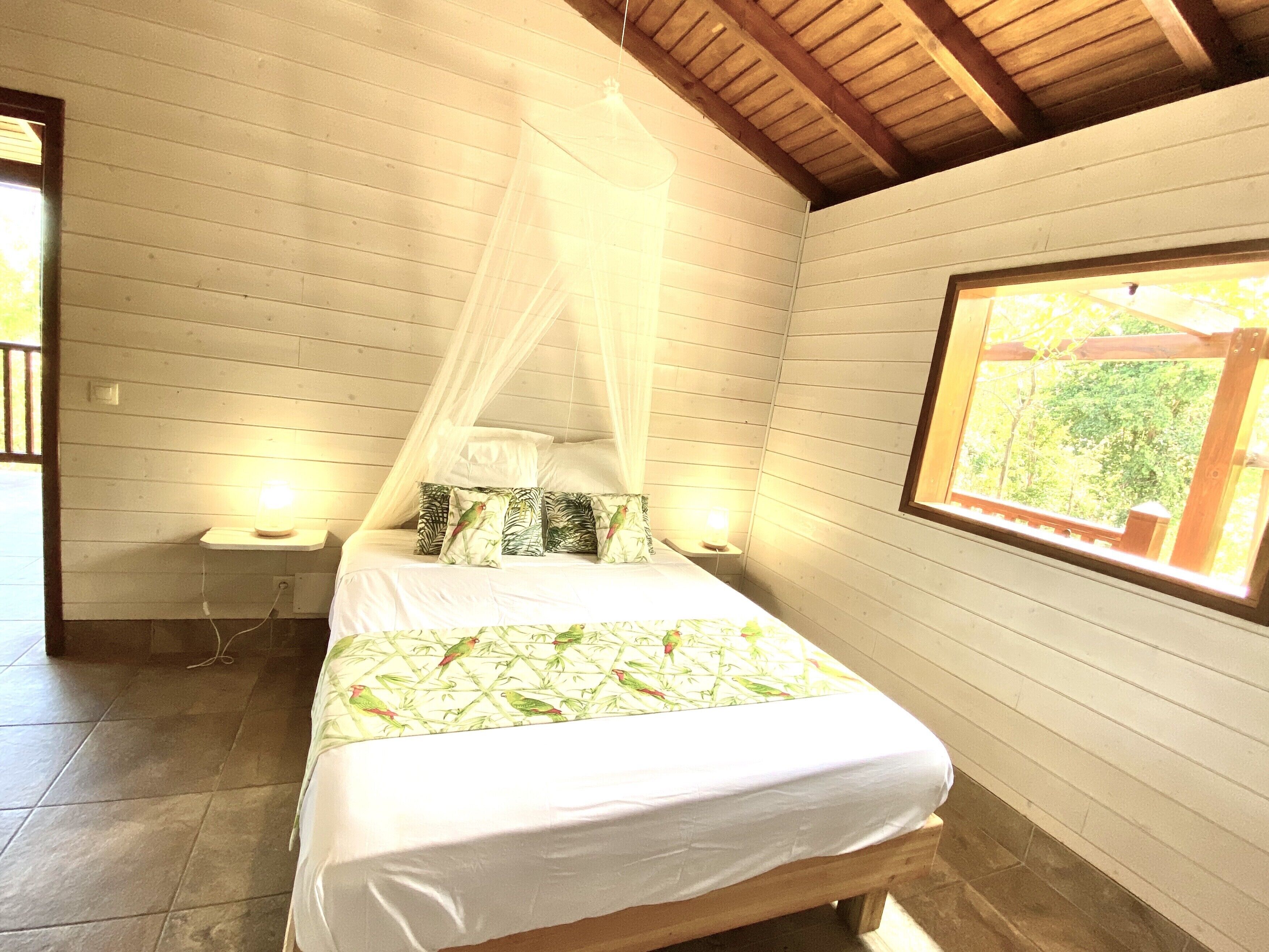Un dormitorio con una cama, dosel, dos mesitas de noche con lámparas y una ventana con vista a follaje.