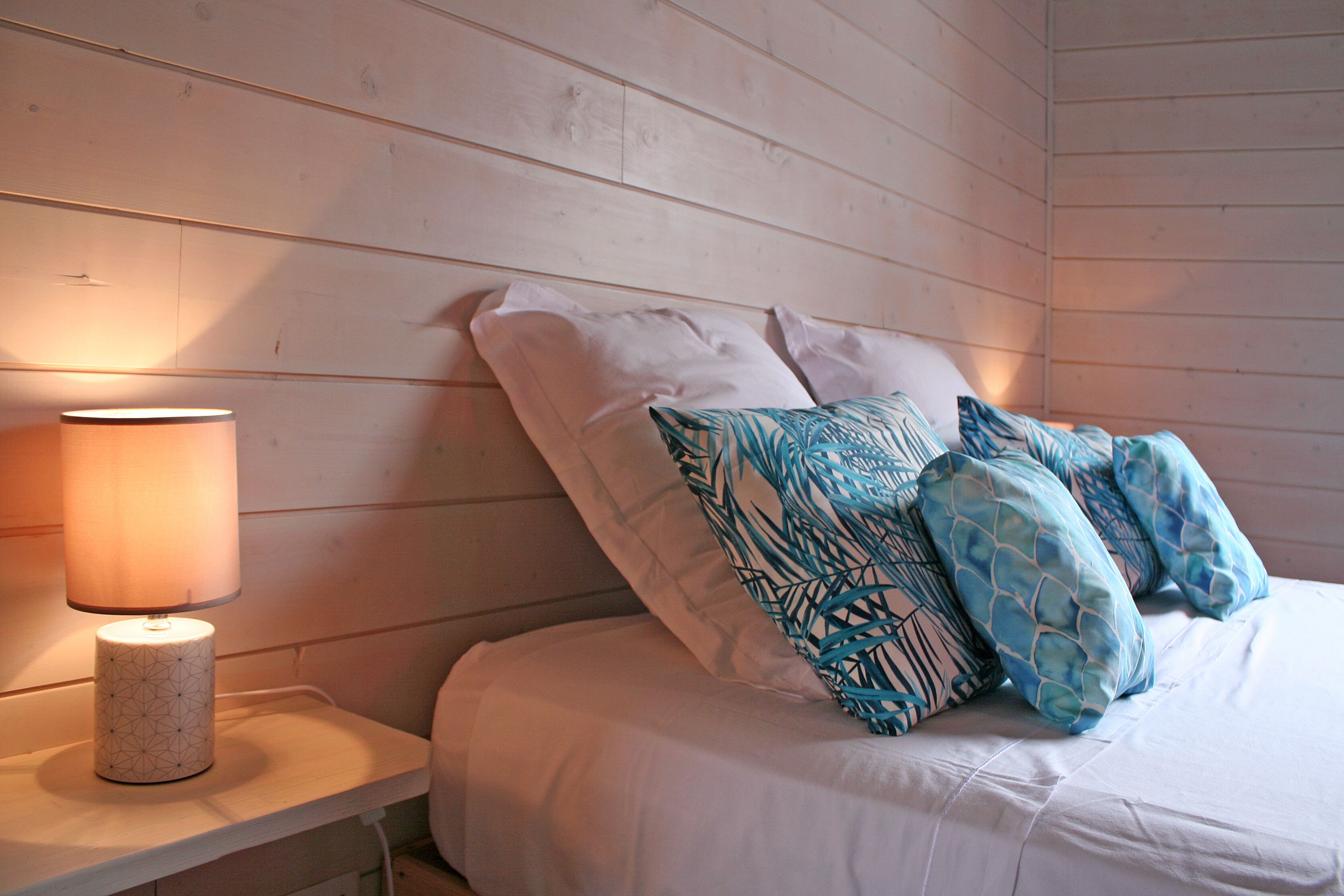 A bedroom with a wooden headboard, a bedside table with a lamp, and a bed with white bedding and patterned pillows.