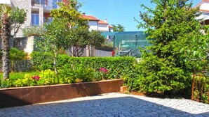 A courtyard with a stone floor, surrounded by greenery and a building in the background.