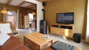 A modern living room with a wooden sofa, a coffee table, a TV, and a kitchen area in the background. - 2017 ecologically renovated, high quality furnished cottage with yard. (Lüneburg)