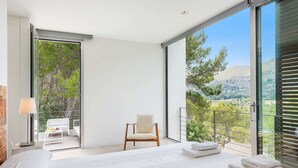 A modern hotel room with a large bed, a chair, and a balcony with a view of trees and hills.