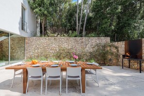 A wooden dining table set for six with white chairs, a stone wall, and a grill in the background.