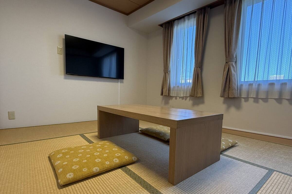 Een kamer met tatami vloerbedekking, een lage houten tafel, een aan de muur bevestigde televisie en een raam met gordijnen.