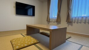 A room with tatami flooring, a wooden table, a television mounted on the wall, and a window with curtains.