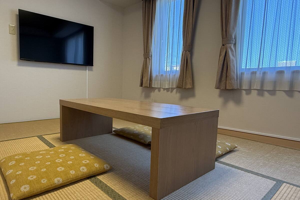 Een kamer met tatami vloerbedekking, een houten tafel, een aan de muur bevestigde televisie en een raam met gordijnen.