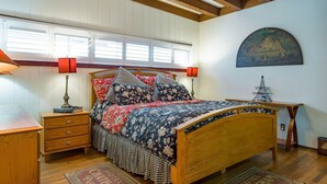 A bedroom with a wooden bed, two bedside tables, a nightstand, a window with blinds, a wall-mounted lamp, and a small table with a lamp.