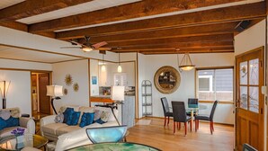 A spacious living room with a high ceiling, wooden beams, a sofa with blue cushions, a glass coffee table, and a dining area with a small table and chairs.