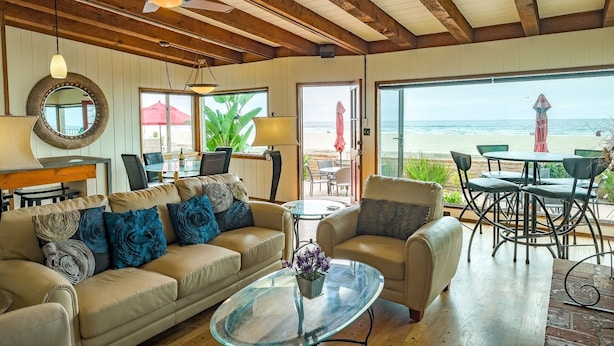 A cozy living room with a sofa, coffee table, and dining area. A large window offers a view of the beach.