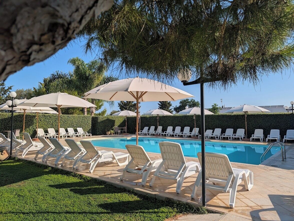 Zona de piscina con sillas reclinables blancas y sombrillas, rodeada de vegetación y un cielo despejado.