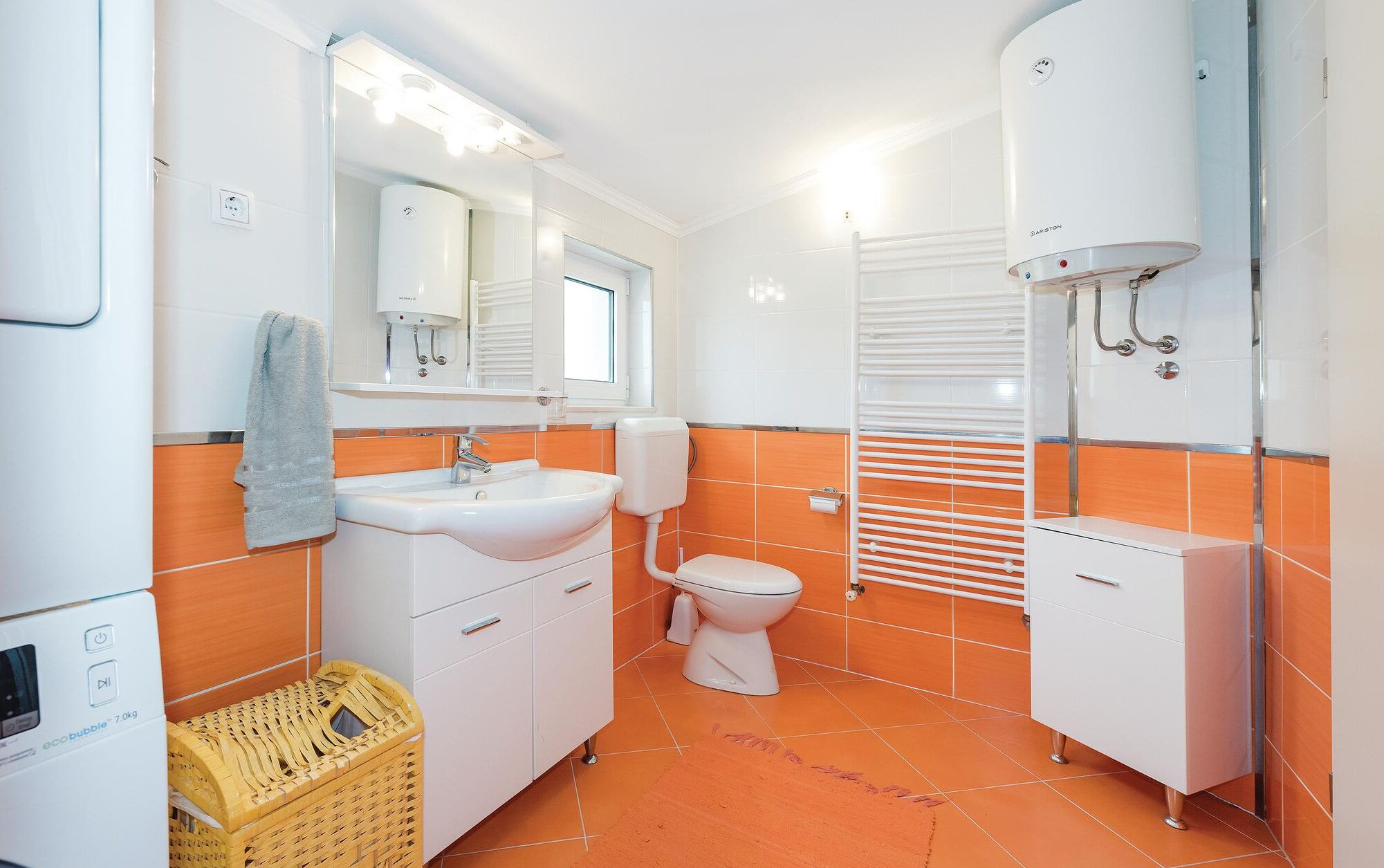 A bathroom with a sink, toilet, and shower. There is a mirror above the sink, and a window above the toilet. The walls are orange, and there is a white cabinet next to the toilet.