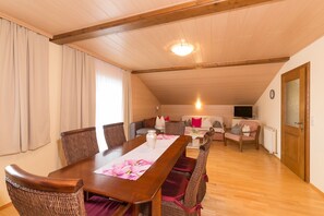Ein gemütlicher Essbereich mit Holztisch und Korbstühlen, einer Sofaecke und einem Fernseher im Hintergrund.
