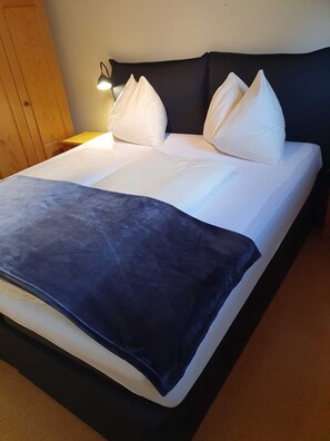 Una cama bien tendida con sábanas blancas y una manta azul oscuro, una lámpara de noche y un armario de madera.
