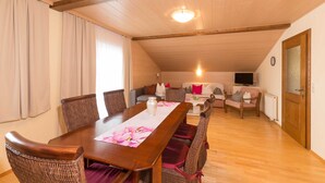 Zona pranzo con tavolo in legno, sedie in vimini e vista su un soggiorno con divano e televisione.