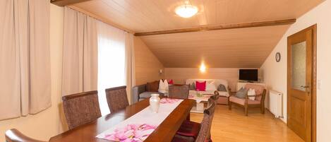 Un comedor con mesa de madera, sillas de mimbre y vista a una sala de estar con sofá y televisor.