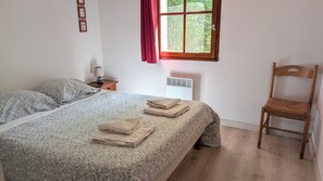 A bedroom with a bed, a chair, a nightstand, a lamp, a window with curtains, and a wall-mounted air vent.
