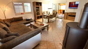 A living room with a sectional sofa, a dining table with chairs, a TV, and a wood-burning stove. - Holiday home FLOW DREAM - Holiday home FLOW DREAM (Göhren-Lebbin)