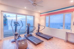 Une salle de sport équipée de tapis de course et d’appareils elliptiques, de grandes fenêtres offrant une vue sur la mer et d’un ventilateur de plafond.