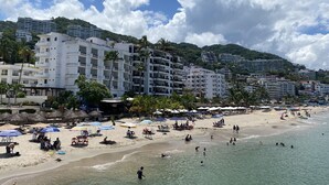 Ett semesterparadis vid stranden med flera vita byggnader, palmer och en sandstrand med parasoller och folk.