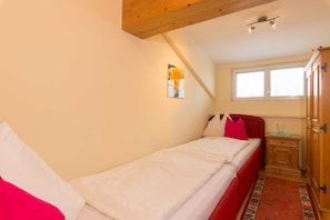 A single bed with white bedding and red headboard, a wooden nightstand with a glass, a small window, and a wall-mounted light fixture.